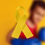 Childhood cancer Awareness graphic: blurry photo of a young boy with boxing gloves and a yellow ribbon in the forefront