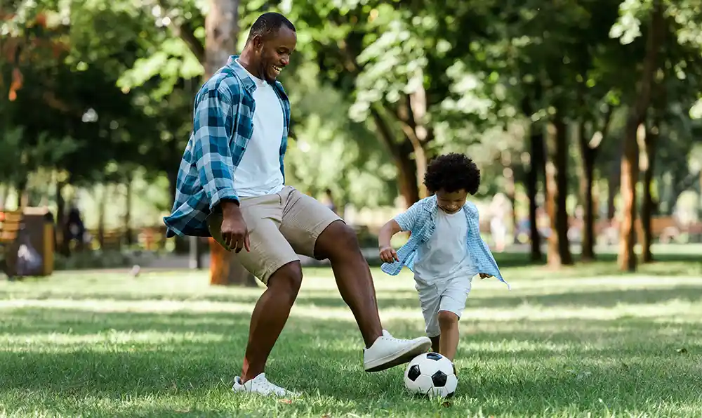 A man enjoys kicking a soccer ball around with his son at a park
