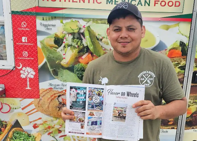 Cachita’s Kitchen Owner Jose Cruz in front of his food truck holding a copy of his article in Mount Pleasant Magazine.