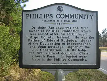 Phillips Community Historical Marker, reads: (Continued from other side) Dr. John Rutledge was the first owner of Phillips Plantation which was named after his birthplace in Phillips County, Ireland. He was the father of Edward Rutledge, signer of the Declaration of Independence, and John Rutledge, signer of the U.S. Constitution. Dr. Rutledge, the first medical doctor in Christ Church Parish, is buried near here in the Phillips Community. Erected in 2002.