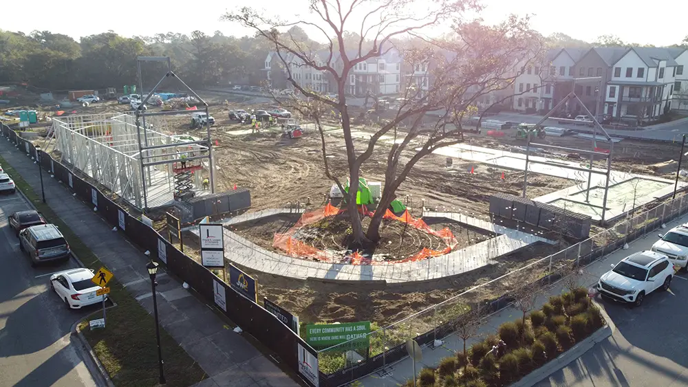 An aerial construction view of the Gather MTP community hub campus in Midtown Mount Pleasant, SC, featuring a preserved grand oak tree surrounded by safety fencing amidst building framing.